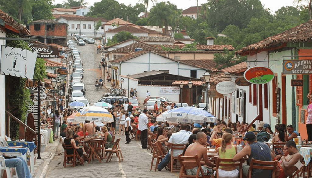 Rua movimentada do centro histórico de Pirenópolis com mesas de restaurantes e guarda-sóis na calçada e carros estacionados, mostrando a vida noturna e o comércio.