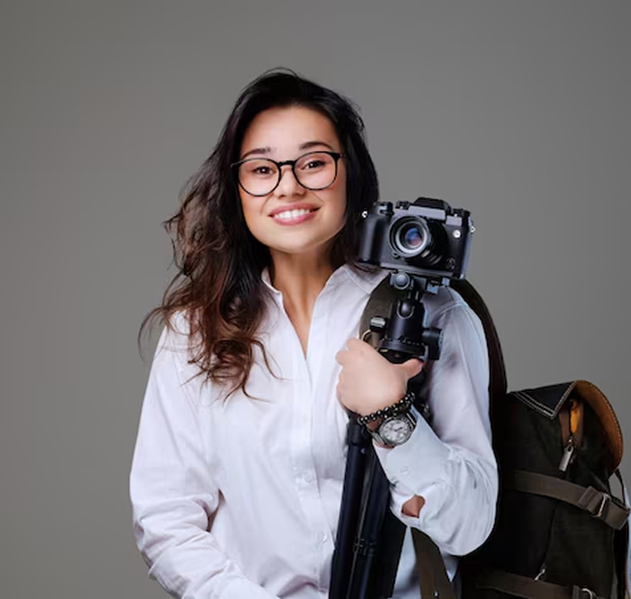 Mulher sorridente com óculos e camisa branca, segurando uma câmera fotográfica e tripé no ombro, com uma mochila de viagem. Representa um fotógrafo de viagem ou guia de turismo.