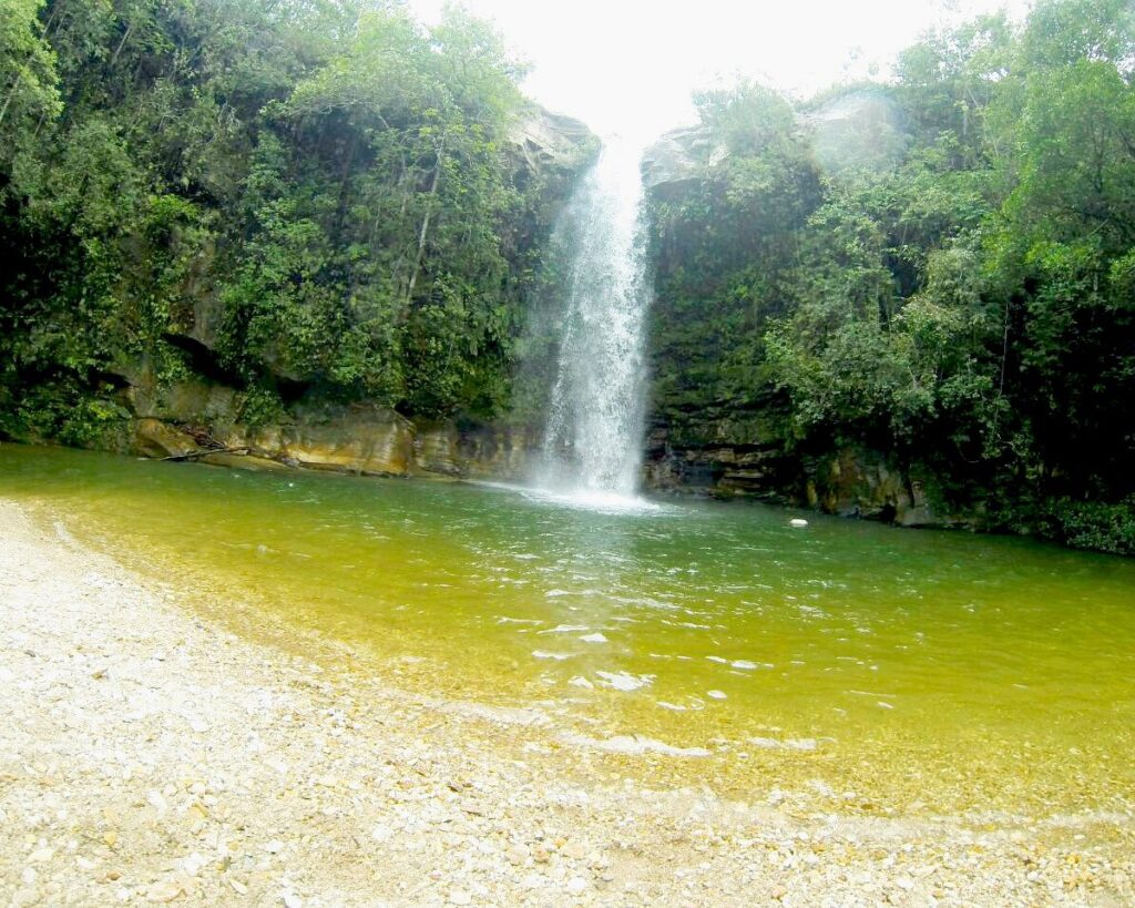 Cachoeira de queda d'água em meio à mata verde, com uma piscina natural de água esverdeada e uma pequena faixa de areia. Ponto de ecoturismo e atração natural de Pirenópolis.