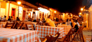 Mesas de restaurante com toalhas xadrez vermelho e branco e clientes sentados na rua de paralelepípedos, iluminadas por luzes quentes à noite, no centro histórico de Pirenópolis.