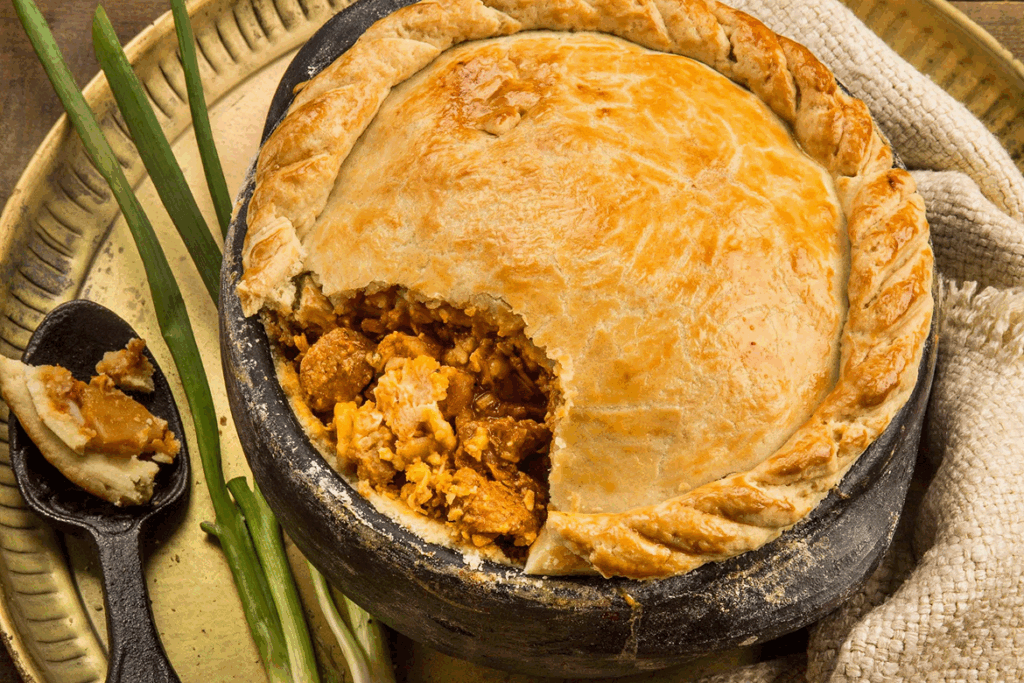 Empadão goiano ou torta salgada rústica na panela de barro, com a casca dourada e recheio de carne e açafrão à mostra, servido com cebolinha verde.