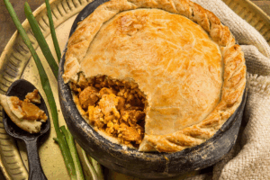 Empadão goiano ou torta salgada rústica na panela de barro, com a casca dourada e recheio de carne e açafrão à mostra, servido com cebolinha verde.