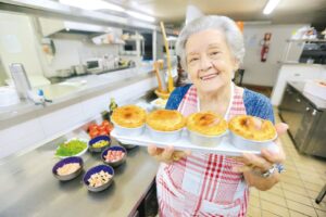 Cozinheira, com avental xadrez, sorrindo e segurando uma bandeja com quatro empadinhas recém-assadas em uma cozinha profissional. Representa a culinária caseira e chefs locais de Pirenópolis.