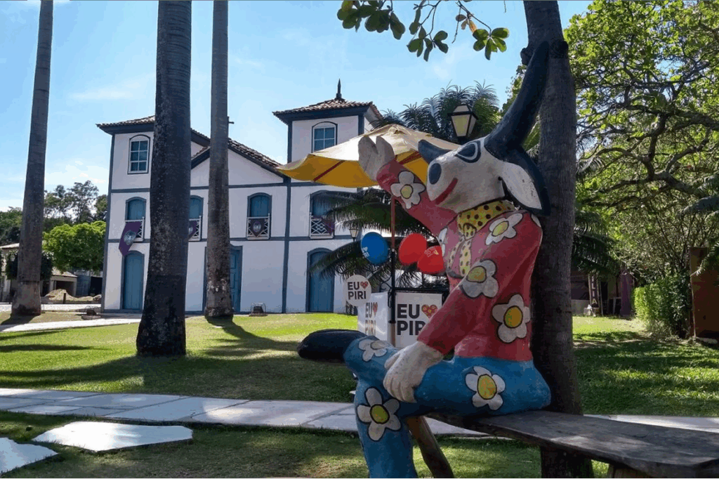 Estátua colorida do Piropiro, personagem folclórico de Pirenópolis, sentado em frente à Casa de Cultura e palmeiras, sob o sol.