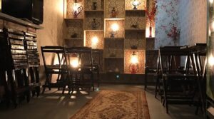 Interior de um bar ou lounge com iluminação baixa e quente, mesas e cadeiras dobráveis de madeira e prateleiras decorativas iluminadas com luminárias rústicas. Representa a vida noturna e os bares temáticos de Pirenópolis.